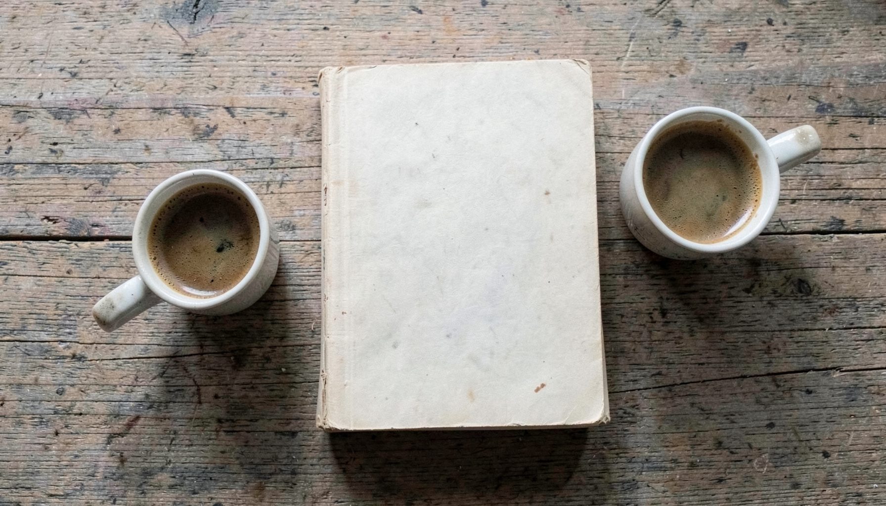 Overhead flat-lay photograph of a worn paperback novel resting on a weathered oak tabletop, beside a half-empty espresso cup, with soft natural light and muted neutral tones